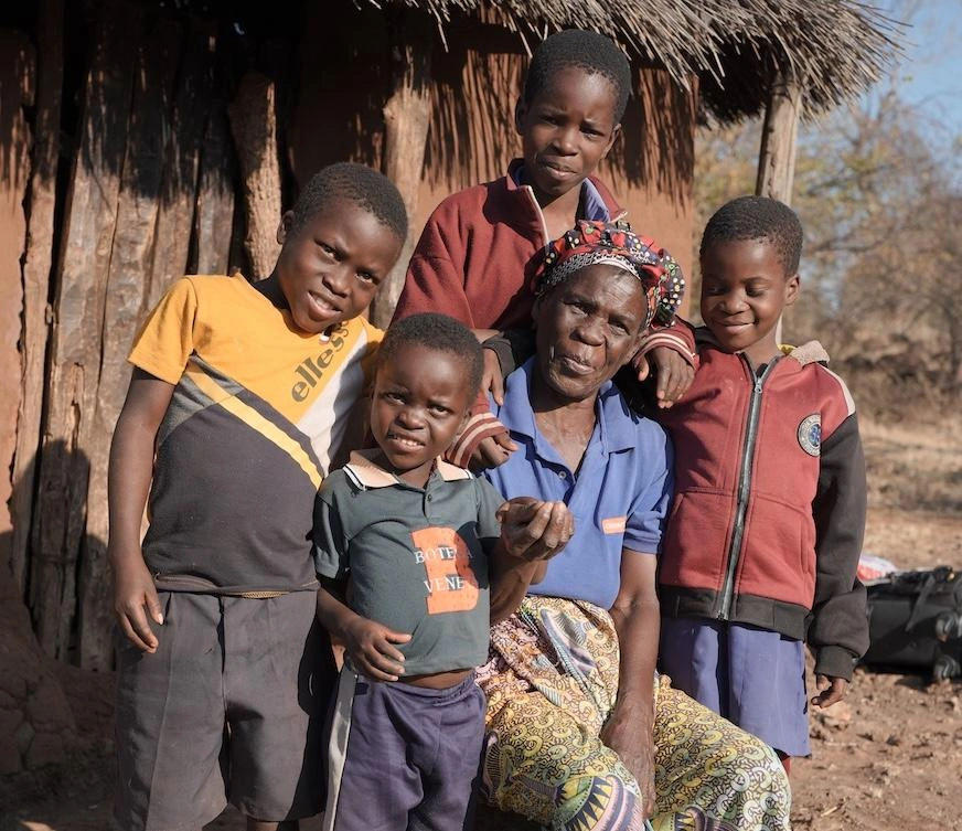 An older woman poses with four children in front of a mud hut.