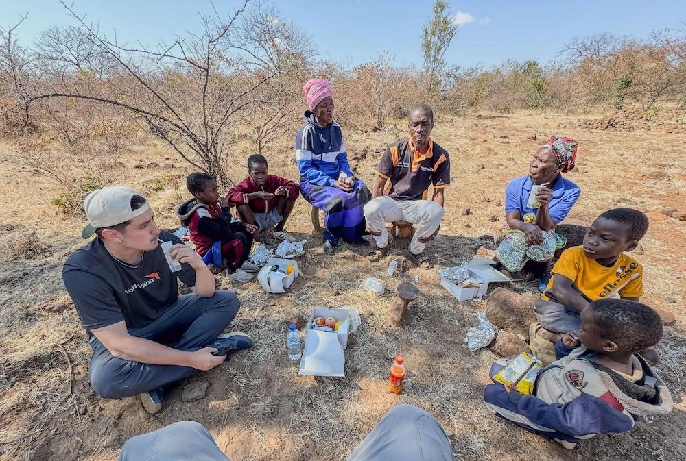 A group of people sit on the ground in a circle eating a packed lunch.