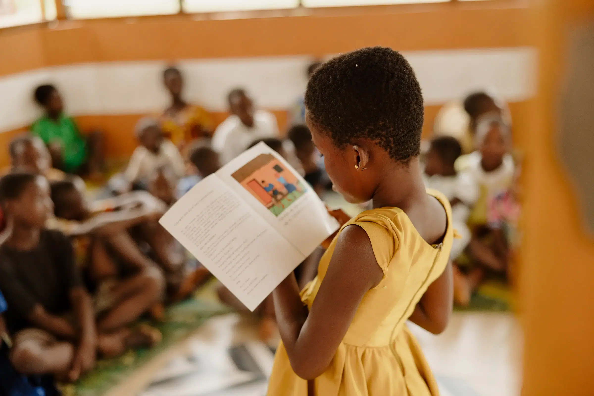 A young girl in a yellow dress is reading a picture book.