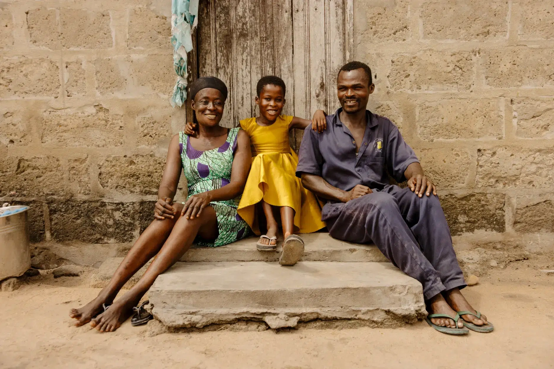 A girl in a yellow dress sits on the entrance to their home between her mom and dad.
