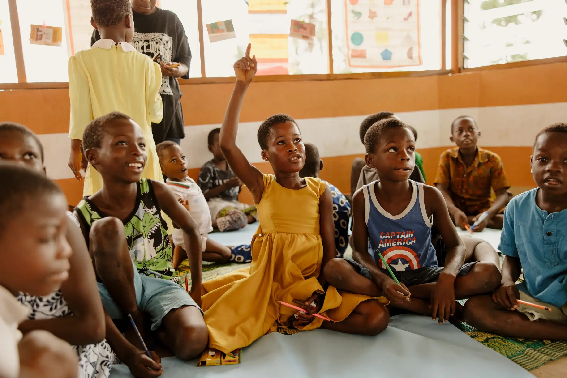A girl in a yellow dress raises her hand as she sits on the classroom floor.