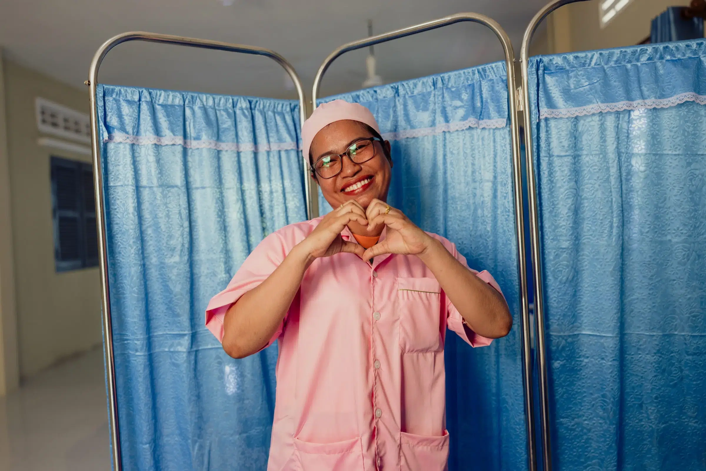 A nurse in pink scrubs stands in front of a blue curtain making a heart shape with her hands.