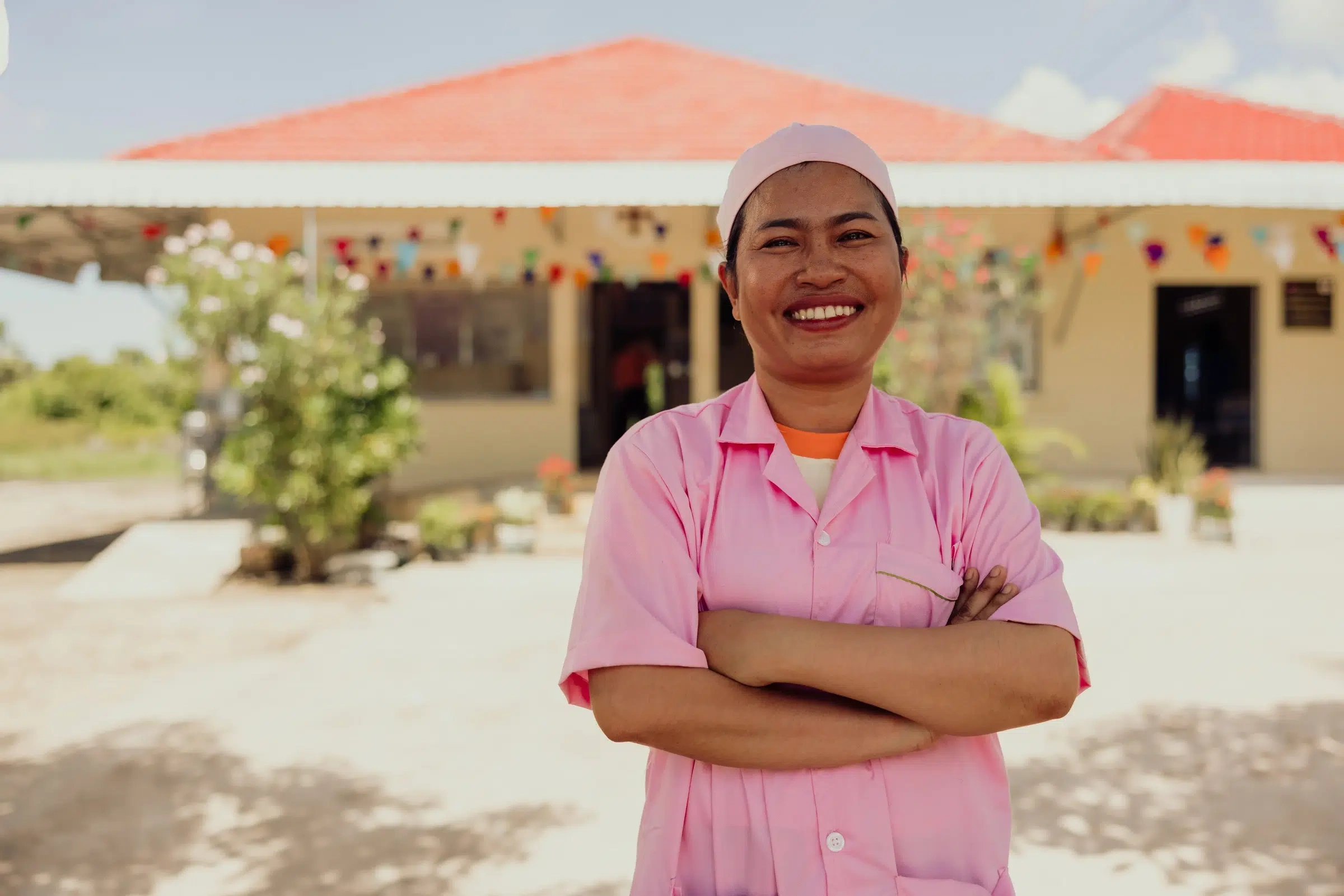 A nurse in pink scrubs with her arms crossed standing outside the clinic where she works.