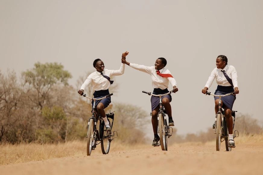 Three girls ride bicycles on a dusty path. Two give each other a high five.