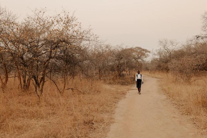 A girl walks a dusty path lined with low-lying scrub and dry grass.
