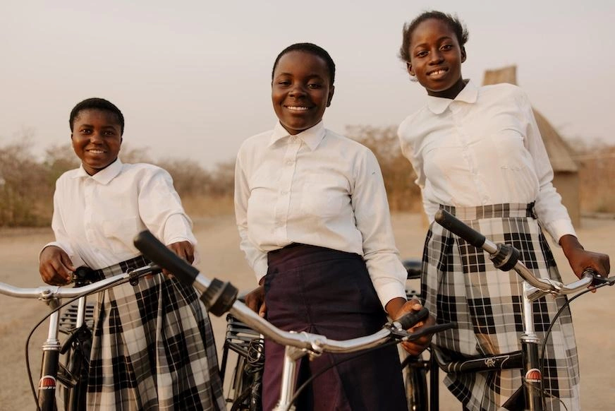 Three teenage girls stand beside shiny bicycles while smiling.