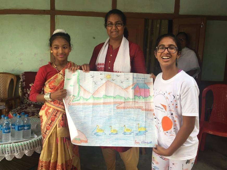 A woman stands smiling between two adolescent girls. All three hold a large landscape drawing.