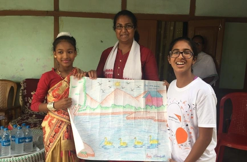 A woman stands smiling between two adolescent girls. All three hold a large landscape drawing.