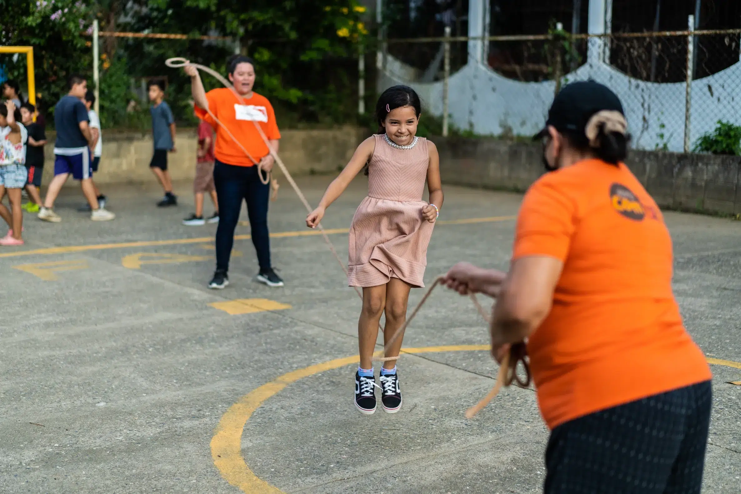 Young girl skipping rope with two women wearing orange shirts.
