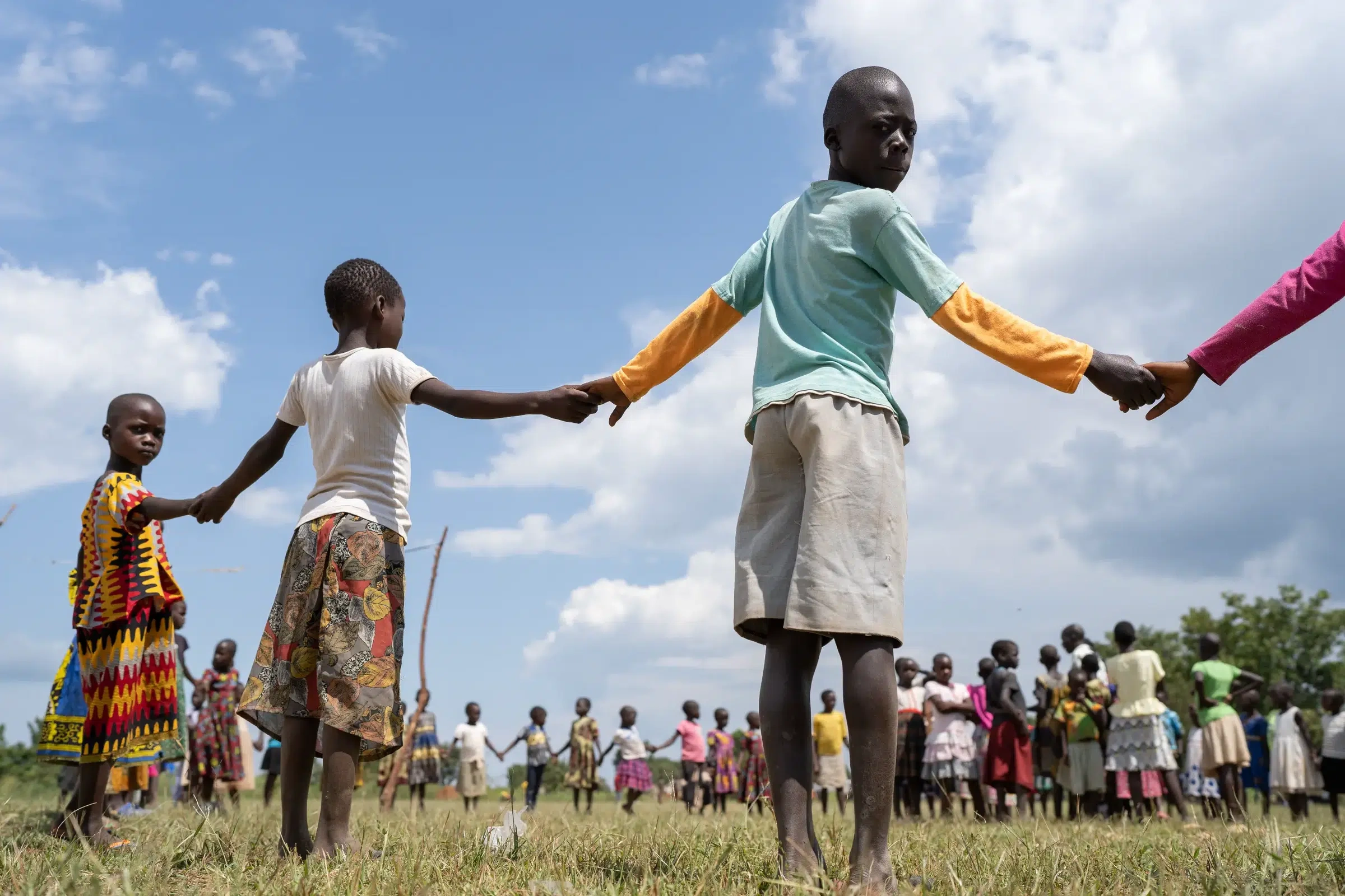 Students in Uganda holding hands in a giant circle under a clear blue sky with white clouds.