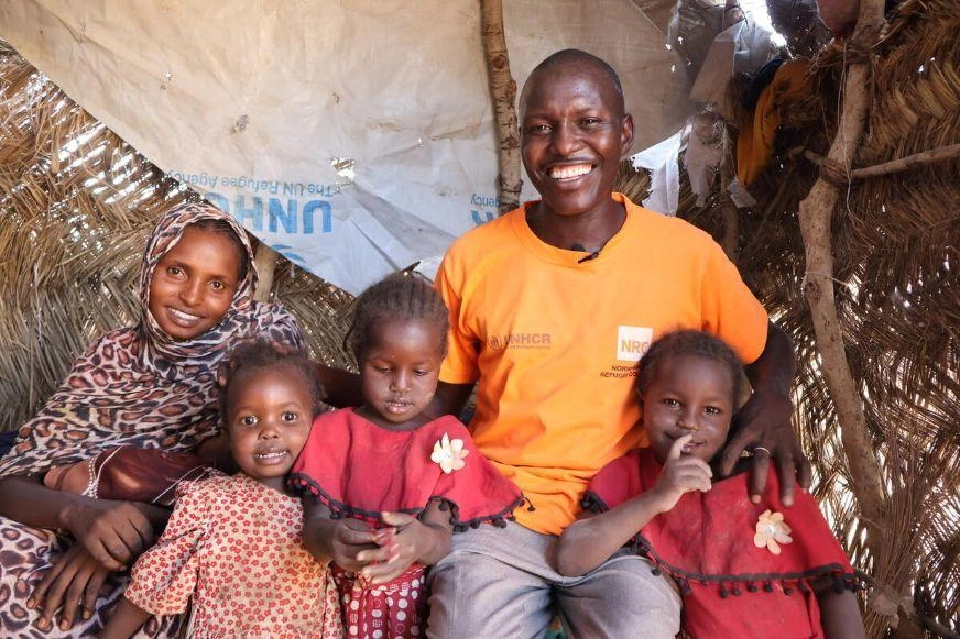Two adults sitting with three children inside a shelter made of woven materials and tarps.