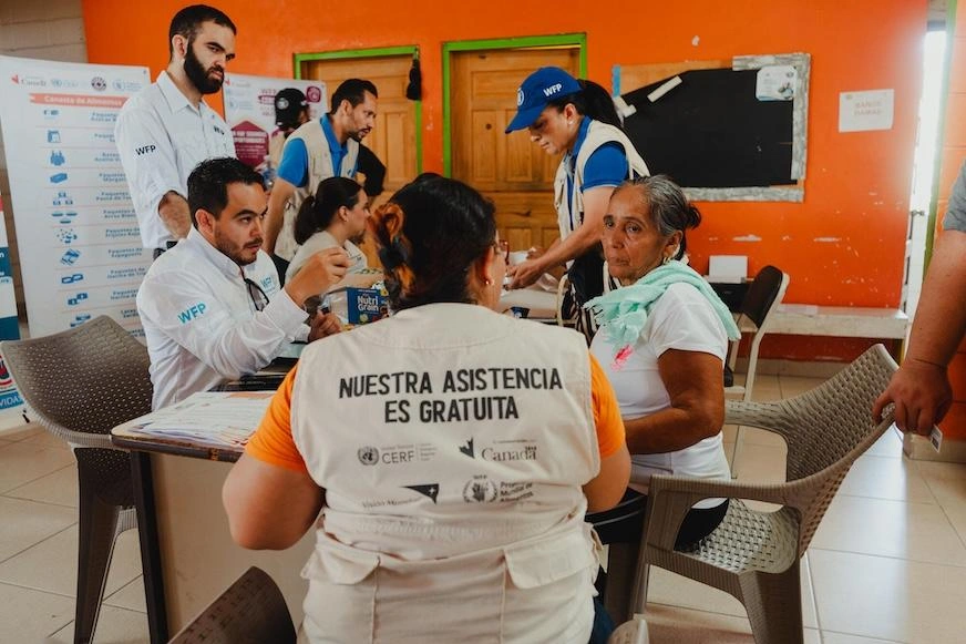 Several people gather around a table in conversation. They wear vests and shirts with World Food Programme branding and World Vision branding.