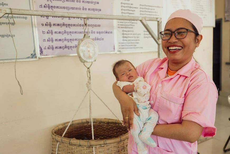 A woman wearing pink scrubs and a scrub cap holds an infant in front of a weigh scale.