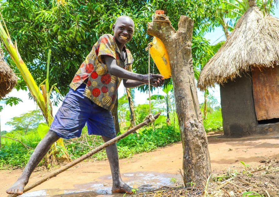 An adolescent boy smiles while demonstrating the use of a modern-day Tippy Tap with a foot pedal.
