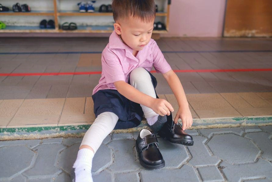 A kindergarten-aged boy sits while putting on shoes with Velcro straps.