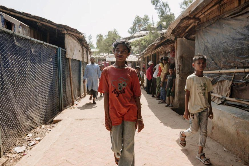 Teenage girl in a red T-shirt walking down a narrow pathway between rows of temporary shelters, with several other people visible in the background.