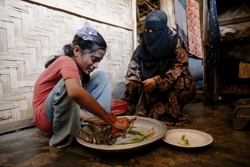 Child preparing vegetables beside a caregiver seated on the floor inside a shelter.