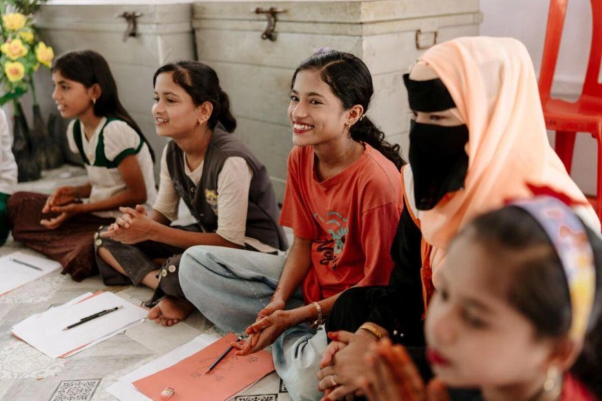Children seated together during a classroom activity, with notebooks and papers on the floor.