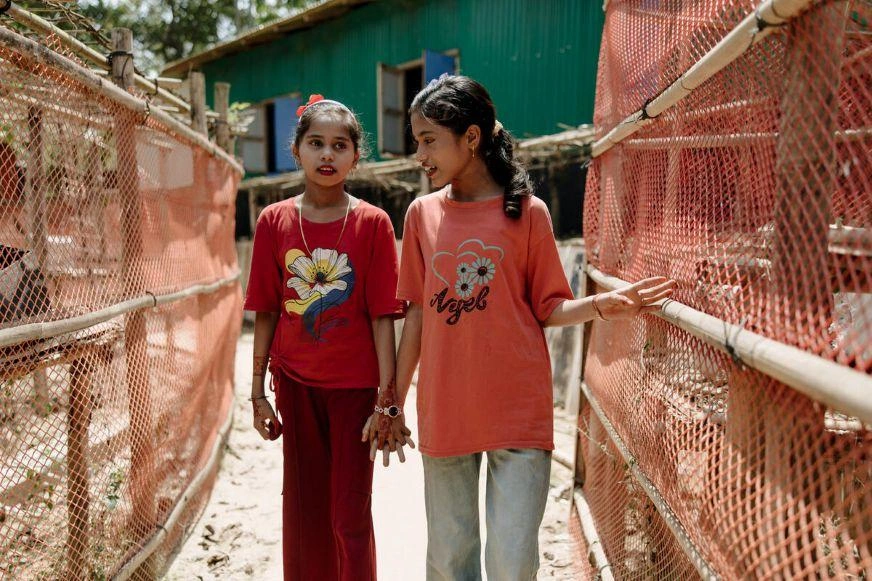 Two young girls walking hand in hand along a narrow pathway between shelters.