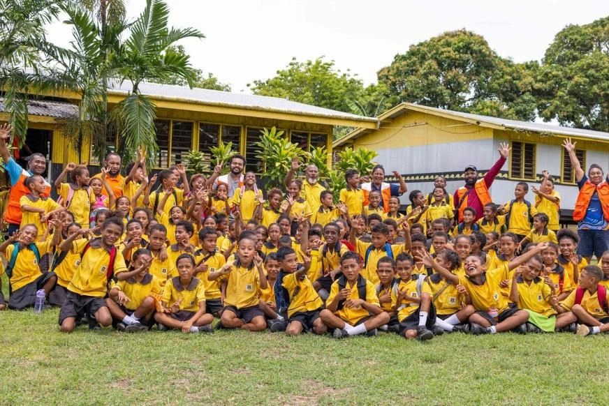 A large group of children and adults in yellow uniforms pose together outside a school building. The group is arranged in rows with trees and greenery visible in the background on a bright day.