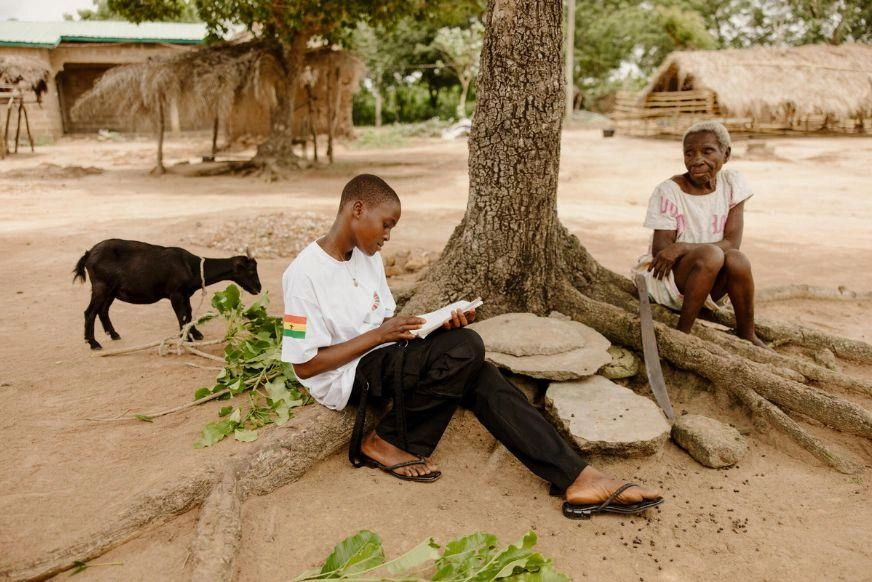 A teenage girl sits under a large tree reading a book. A goat stands behind her and her grandmother sits nearby. The background shows a rural village setting with thatched roof buildings.