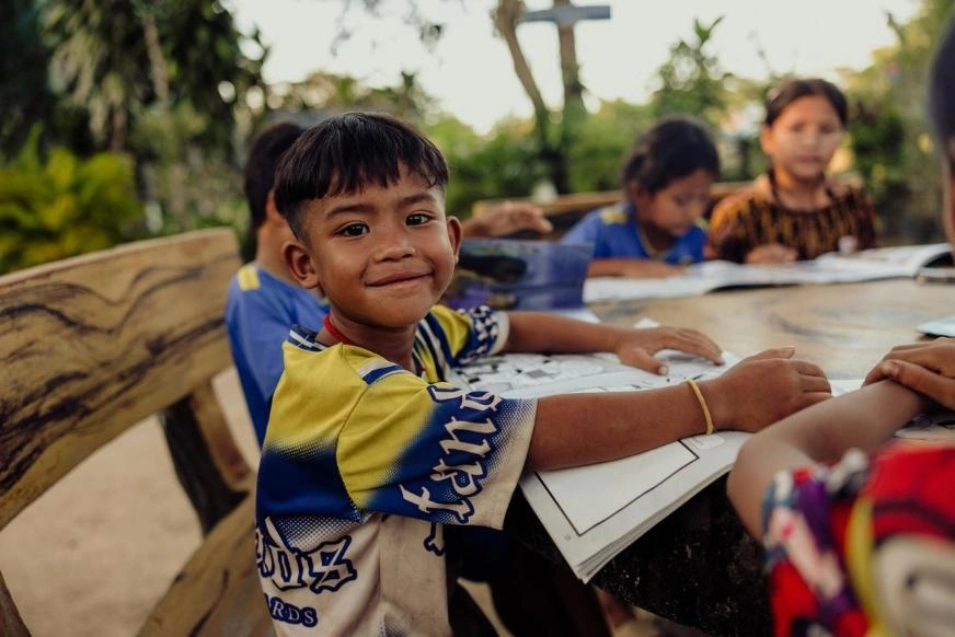 A school-age boy in a blue and yellow shirt sits at a wooden table outdoors, holding a workbook open while participating in a group learning activity. Other students are seated at the table.