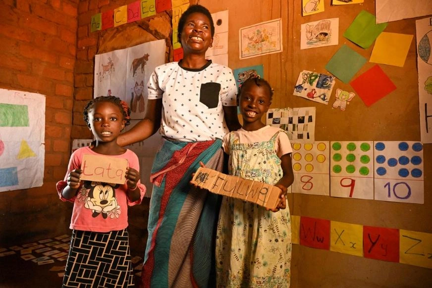 A mother in the middle stands with her two daughters, one on each side, in the learning corner of their home. Colourful drawings, numbers and posters are displayed on the walls behind them.