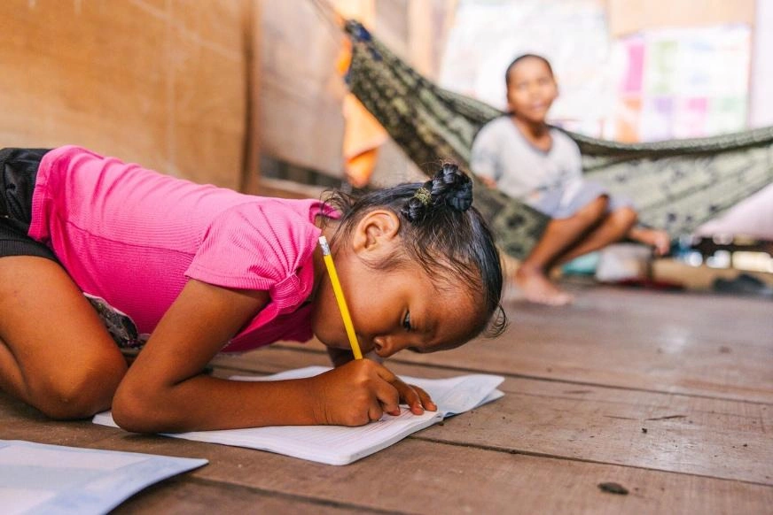 A child in a pink shirt leans over on a wooden floor, concentrating on writing in a notebook with a pencil. In the background, her mother sits near a hammock inside their home.
