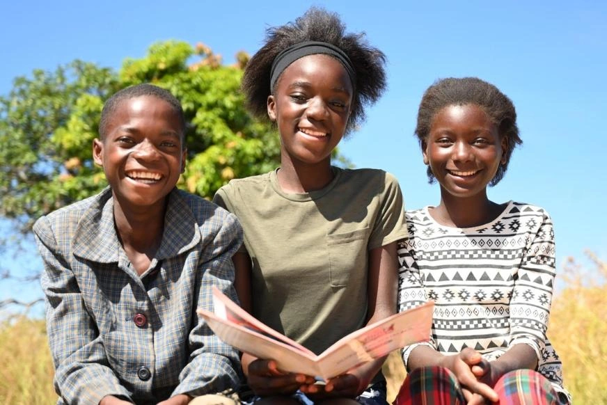 Three young people sit closely together outdoors under a bright blue sky. The girl in the centre holds an open book, with trees and tall grass in the background.