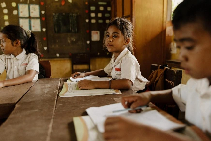 A young girl in a white blouse sits at her school desk as she listens intently to her teacher. Small paper hearts and posters are pinned on a bulletin board behind her.