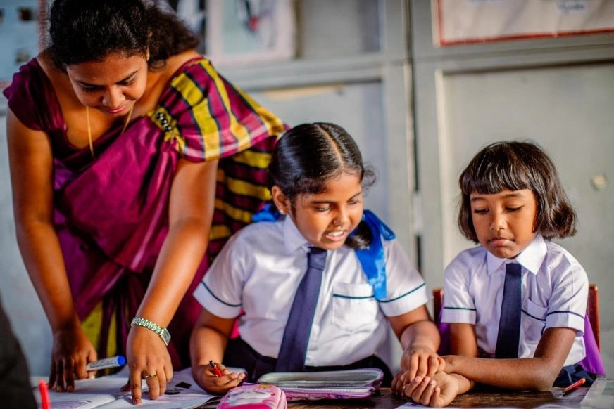 A female teacher leans over a desk to help two students in school uniforms complete schoolwork. One student places something inside the hands of the other.