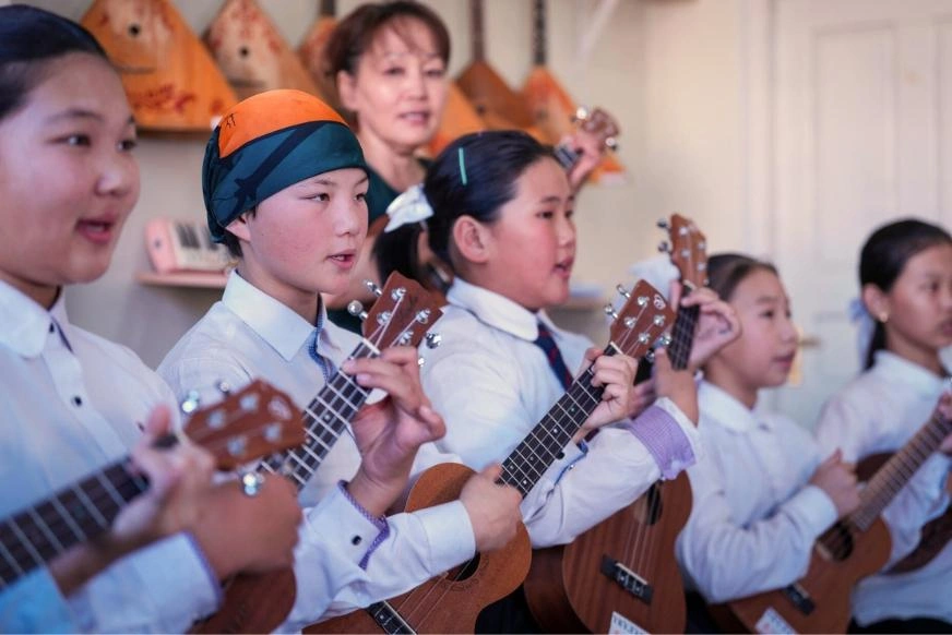 A group of students in school uniforms hold and play ukuleles during a classroom music activity. An instructor stands behind the group and several stringed instruments are visible in the background.
