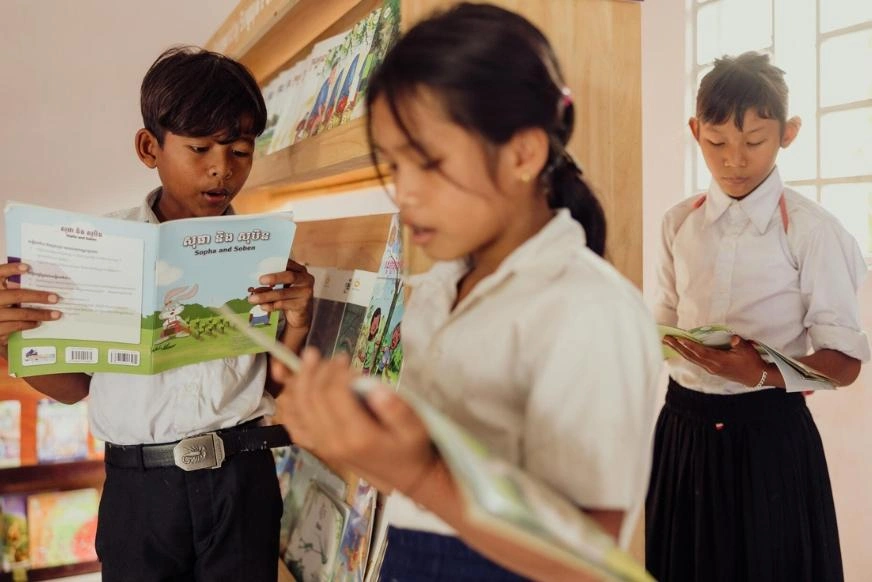 Three school children stand in a school library reading books. The child in the foreground holds an open book while two other children read books behind her. In the background are stocked bookshelves.