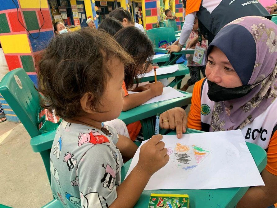 A lady wearing a mask and head scarf crouches beside a preschool-aged child who is drawing with crayons.