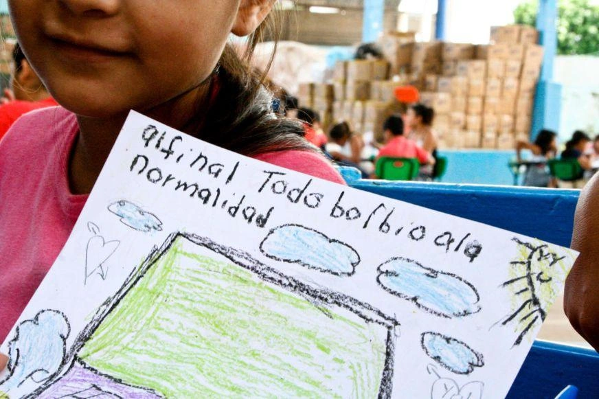 We see the corner of a child’s face as she holds up a crayon drawing showing a roof, clouds and sunshine. She has written a few words in Spanish.