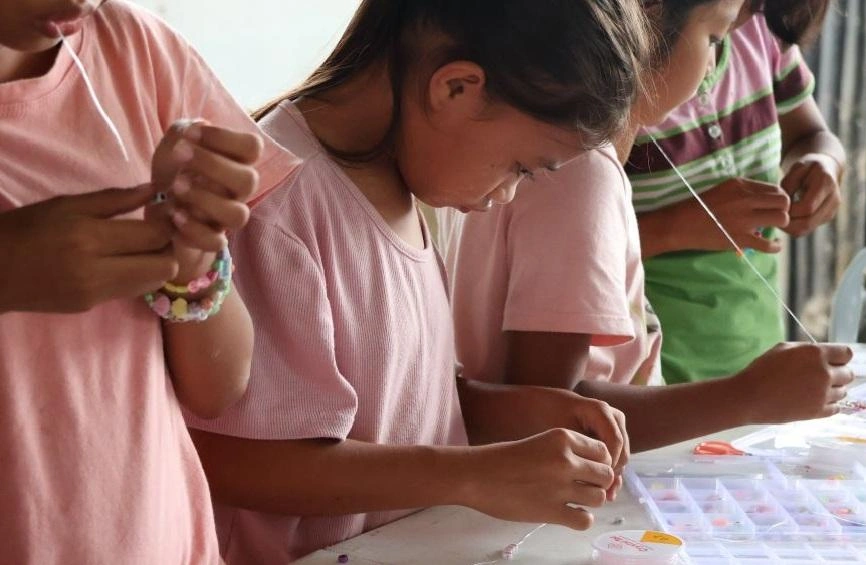 Three children carefully string beads on a string, looking down to focus on their work.