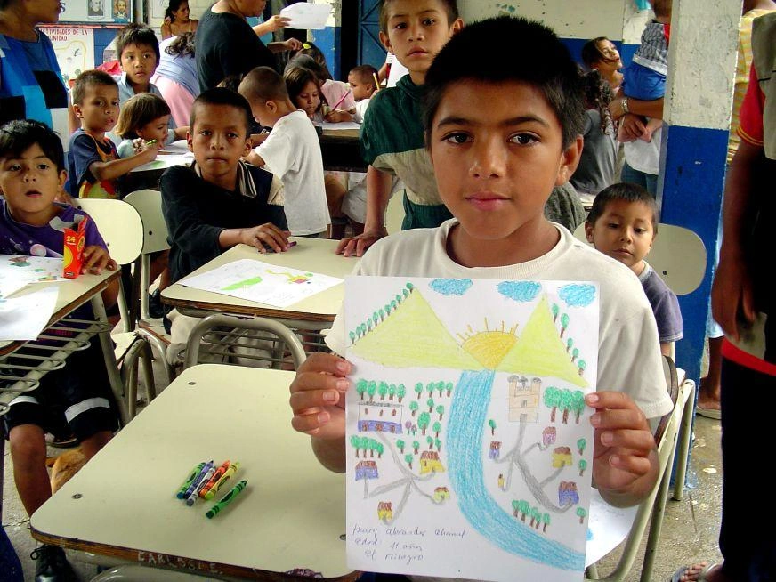 A school-aged boy holds up a drawing of a village with mountains and sunrise behind.
