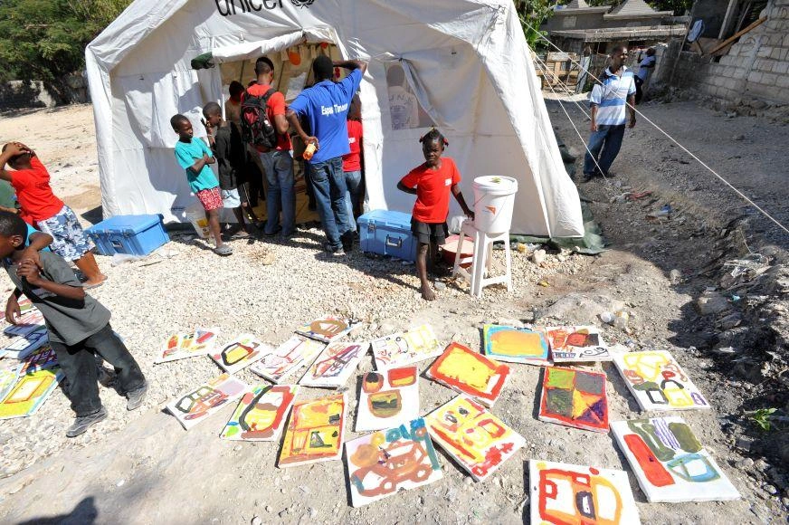 A school-aged child stands outside a white tent, multiple children’s kids’ paintings spread out on the rubble before her.