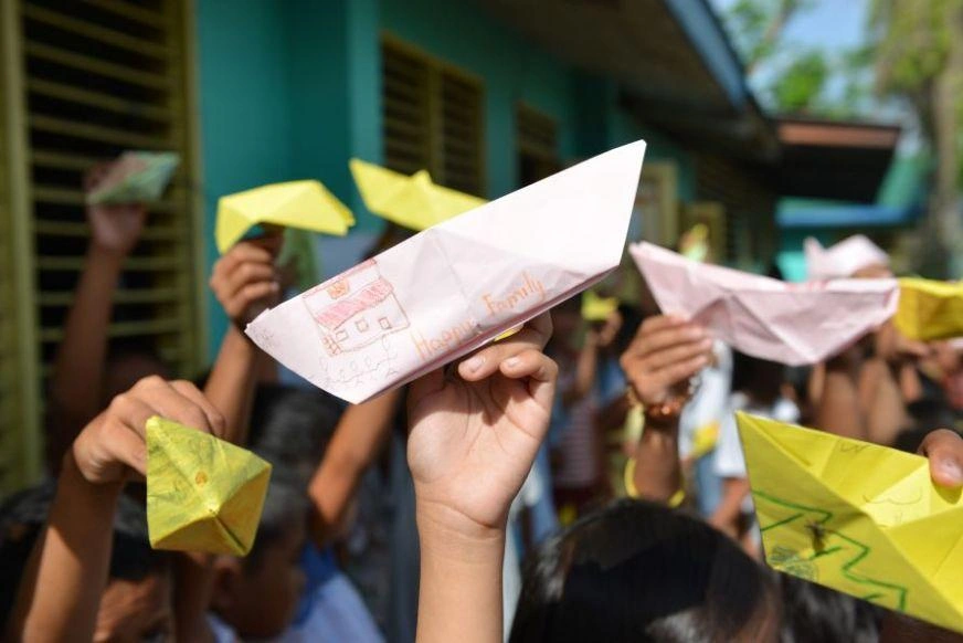 Children’s hands hold up colourful, home-made paper boats covered with pictures they’ve drawn.