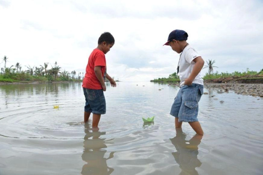 Two school-aged boys paddle into a river, floating their colourful paper boats in the water.