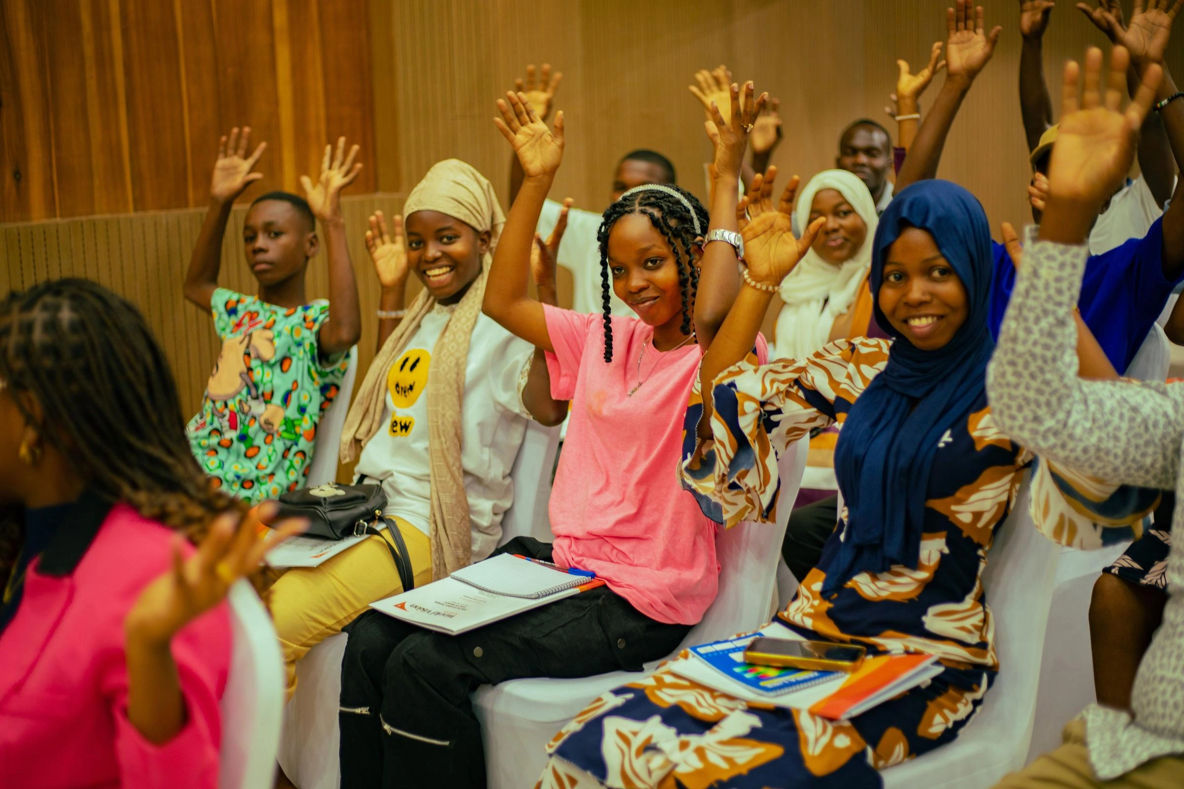 A group of children, sitting, raise their hands above their heads.