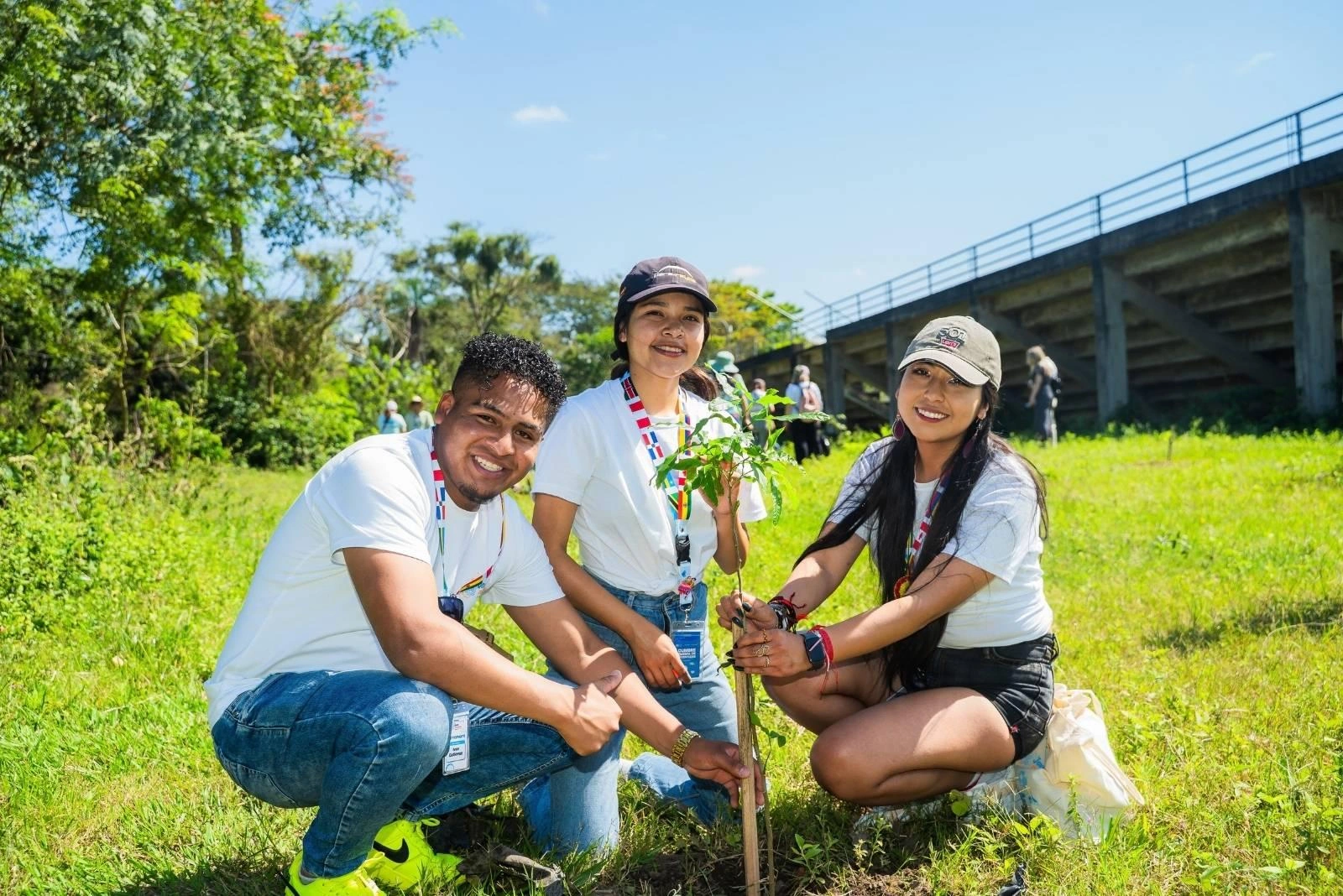 Three young people smile while working together to plant a tree on a sunny day in Bolivia.