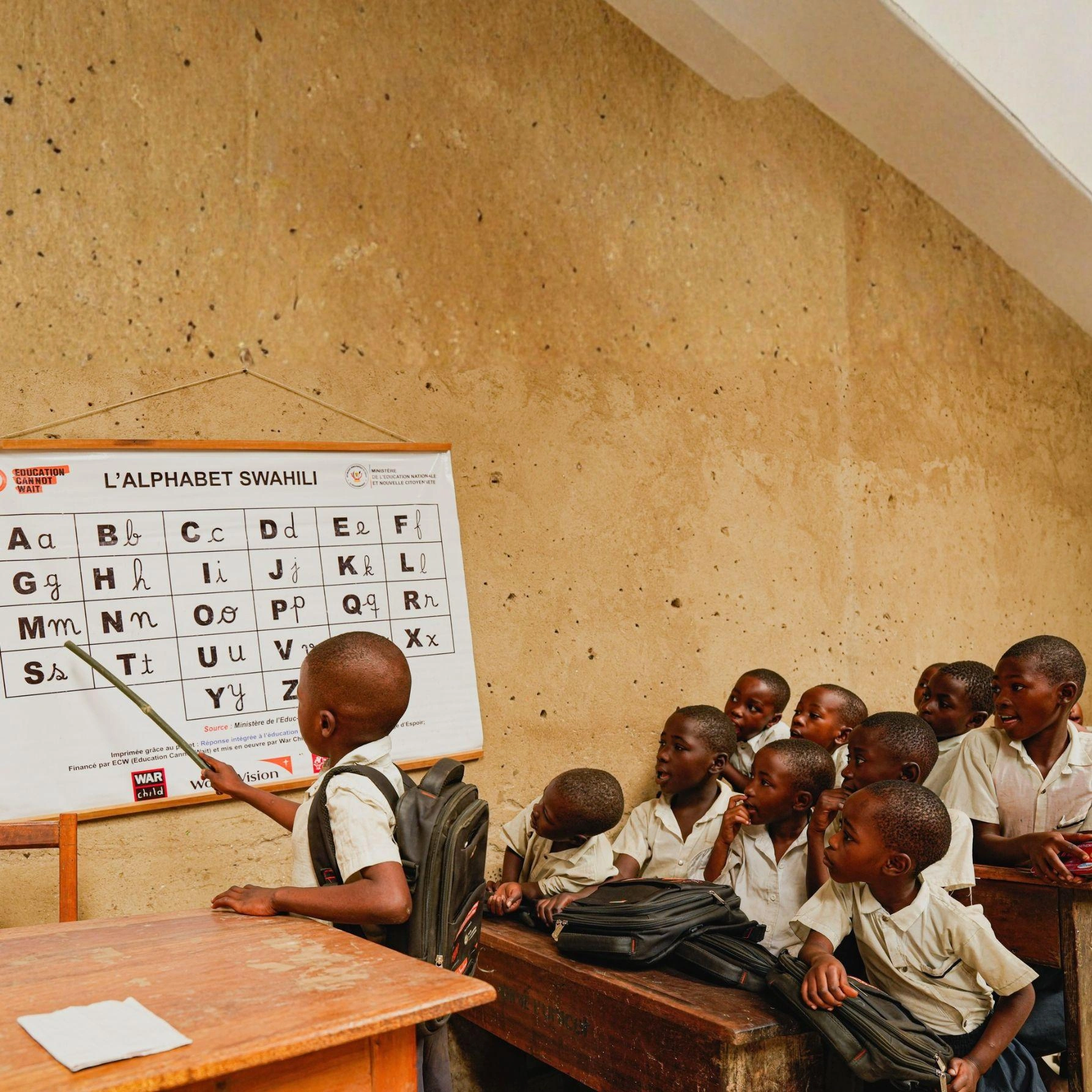 A boy points to the alphabet on a classroom board while his classmates watch attentively.
