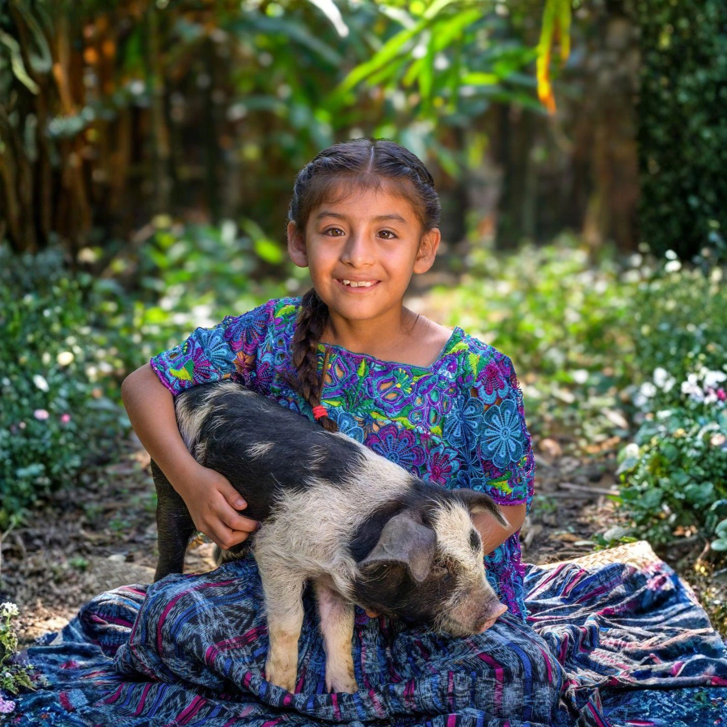 A little girl in a multicoloured dress smiles with joy as she holds her piglet.
