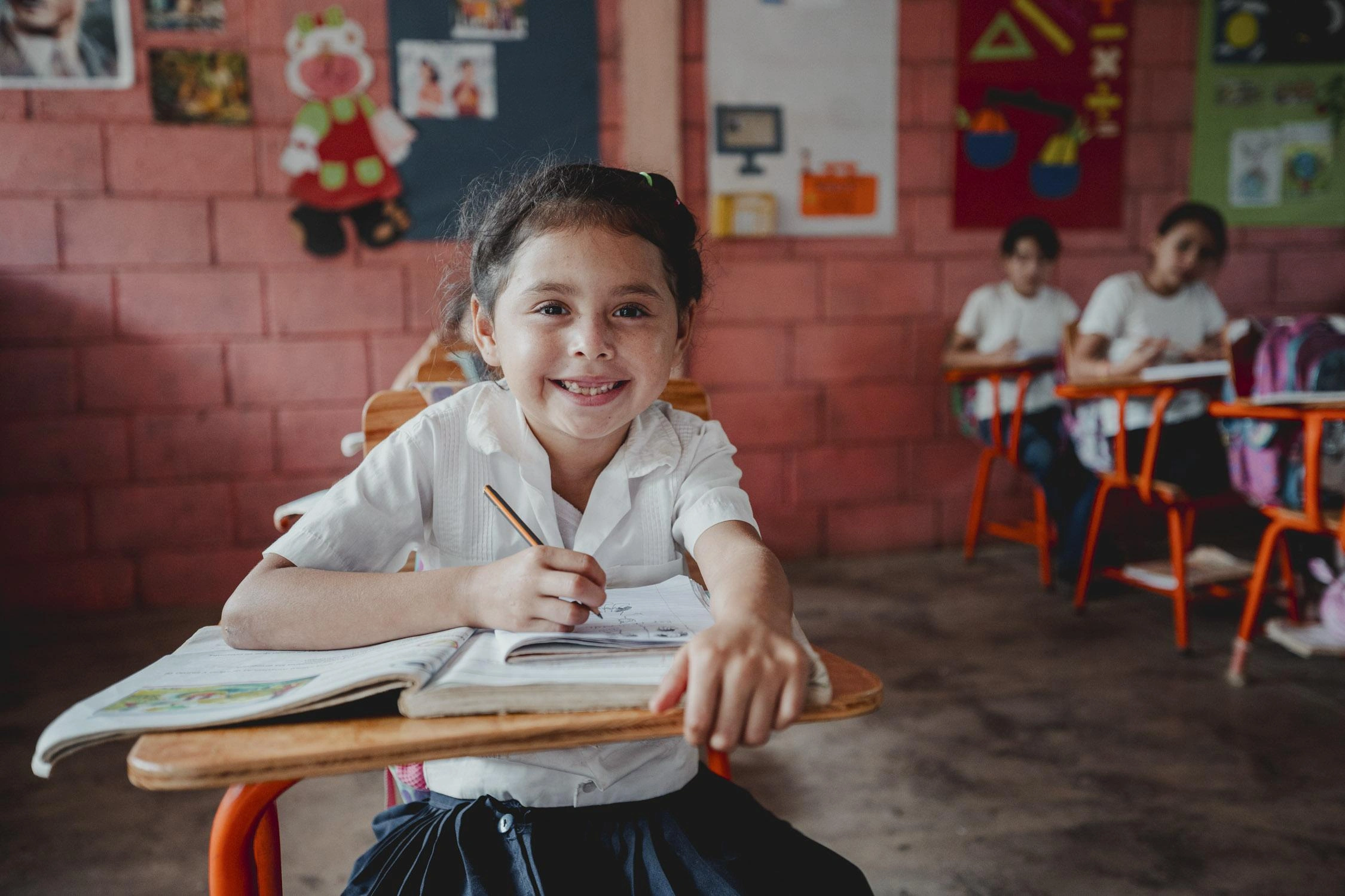 A smiling girl in a white school uniform sits at her desk writing in a notebook.