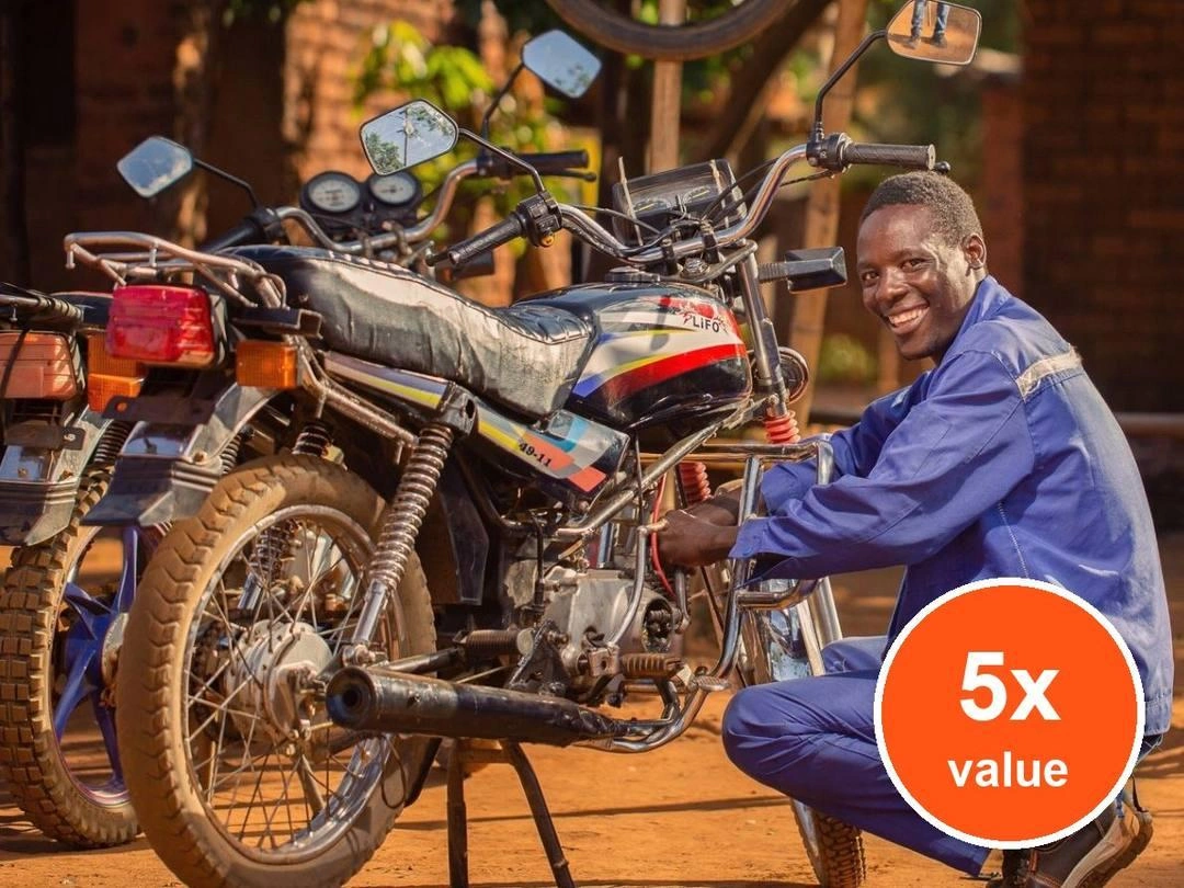 A man in overalls smiles as he squats beside a motorcycle. Badge: 5x value.