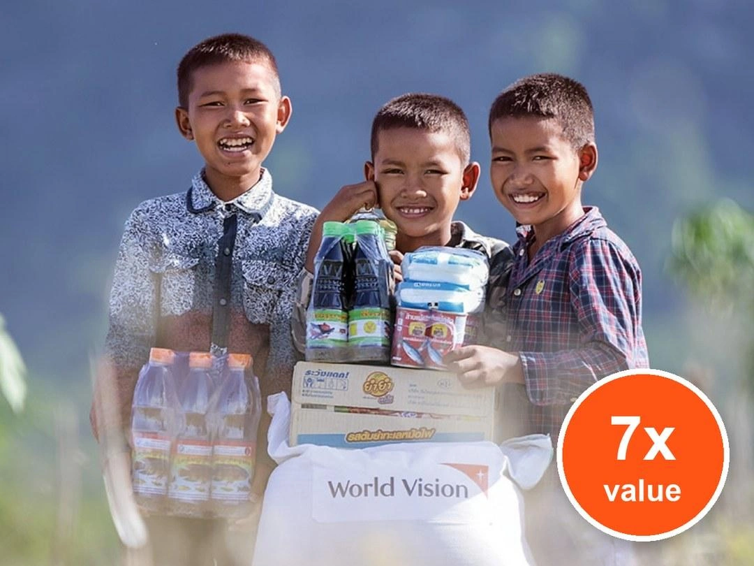 Three boys smile next to each other as they stand behind a pile of food items. Badge: 7x value.