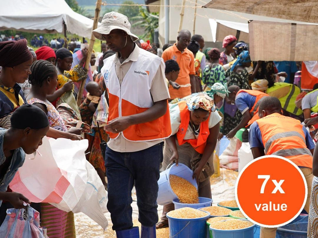 Relief workers in orange safety vests distribute food to community members, scooping grain into buckets and bags at an outdoor aid distribution site. Badge: 7x value.