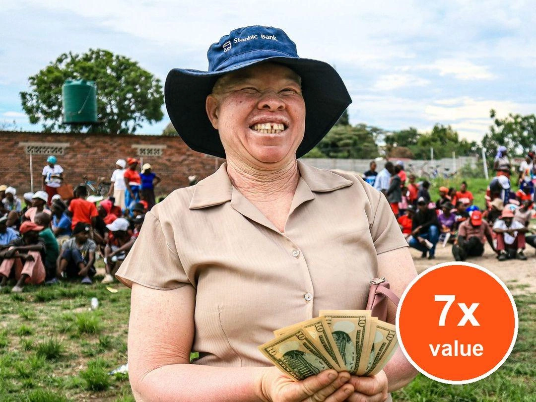 A woman wearing a brown shirt and blue denim sun hat smiles joyfully as she spreads a handful of cash. Badge: 7x value.