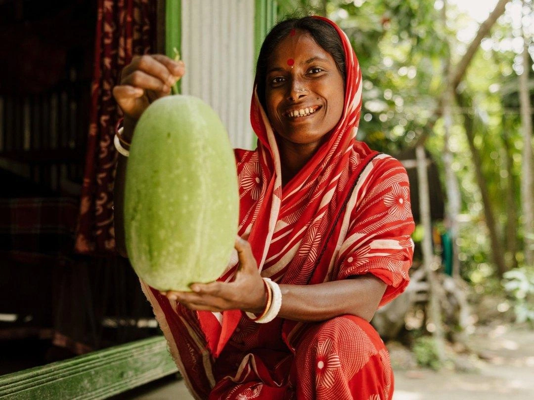 A woman wearing a red and gold sari smiles while holding up her produce.